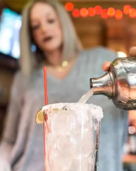 A woman is pouring a clear liquid from a metal cocktail shaker into a glass filled with ice, garnished with a lime wedge and a red straw, in a cozy bar or restaurant setting.